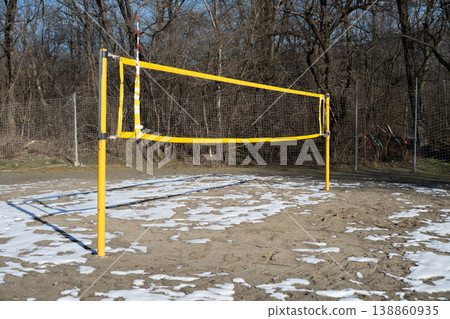Empty outdoor volleyball court with a yellow net set up on sandy ground with patches of melting snow. Concept of outdoor sports, recreation, seasonal change, and solitude. Empty outdoor volleyball court with a yellow net set up on sandy ground with patches of melting snow. Concept of outdoor sports, recreation, seasonal change, and solitude. 138860935