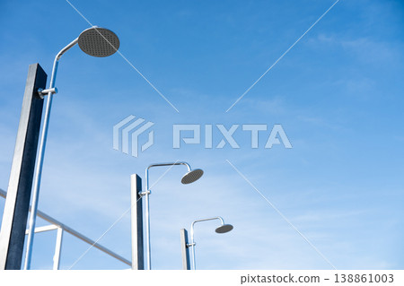 Row of outdoor public showers installed on metal poles against a bright blue sky. Minimalistic low-angle view with large copy space, symbolizing summer, beach facilities, hygiene, refreshment, and Row of outdoor public showers installed on metal poles against a bright blue sky. Minimalistic low-angle view with large copy space, symbolizing summer, beach facilities, hygiene, refreshment, and 138861003