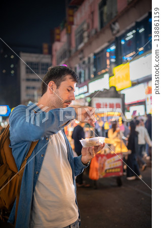 Man Enjoying Street Food at Vibrant Night Market in China, Surrounded by Colorful Neon Lights and Bustling Atmosphere Man Enjoying Street Food at Vibrant Night Market in China, Surrounded by Colorful Neon Lights and Bustling Atmosphere 138861159