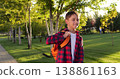 Joyful teenager walking in sunny park with orange backpack, smiling. Youth lifestyle, casual outdoor activity, summer, leisure, calm and happy everyday moment. Teenage schoolboy at sunset or sunrise. 138861163