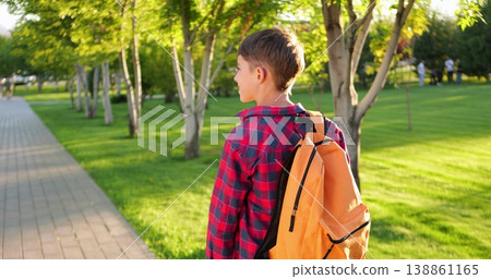 Happy child boy outdoors walks along park path at sunset. Schoolboy boy with backpack smiles. Happy child goes to school. Joyful kid outdoors enjoying nature, golden sunlight, and peaceful walk. 138861165