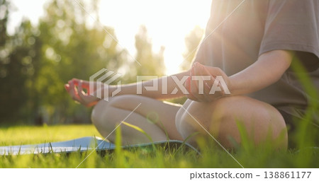 Young woman practices yoga meditation outdoors in park during golden hour. Girl closeup sits in lotus pose green grass sunset. Female lotus position. Healthy lifestyle harmony wellness deep relaxation Young woman practices yoga meditation outdoors in park during golden hour. Girl closeup sits in lotus pose green grass sunset. Female lotus position. Healthy lifestyle harmony wellness deep relaxation 138861177
