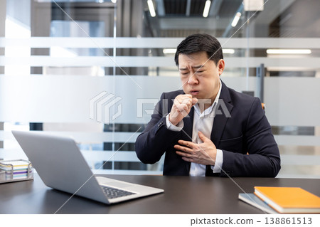 Asian businessman working on laptop experiencing a severe cough and chest discomfort, symbolizing illness, poor health, and the struggle of working while unwell in a professional environment 138861513