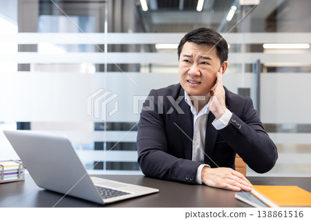 Asian businessman sitting at office desk, suffering from severe earache or tinnitus, covering his aching ear with hand, indicating discomfort and potential health issues during work Asian businessman sitting at office desk, suffering from severe earache or tinnitus, covering his aching ear with hand, indicating discomfort and potential health issues during work 138861516
