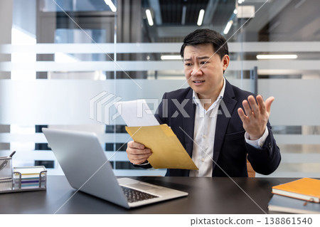 Asian businessman in suit at desk, reacting with worry and hand gestures while opening an envelope and reading a troubling document beside his laptop in a modern office 138861540