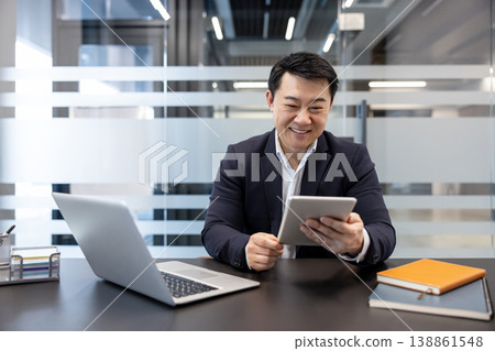 Asian businessman smiling and using a digital tablet at his desk in a modern office, surrounded by technology like a laptop and notebooks, representing productivity and professional communication 138861548