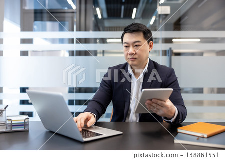 Asian businessman working in a corporate office. Simultaneously typing on a laptop and holding a digital tablet. Demonstrating multitasking and technological integration in a professional environment 138861551