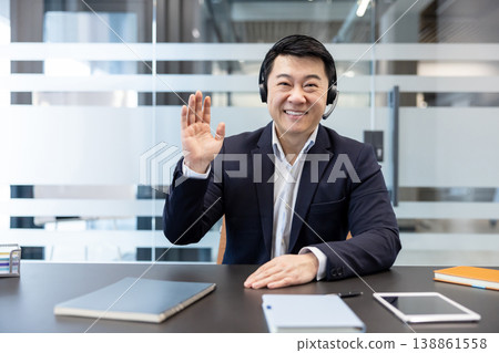 Asian businessman wearing a headset and smiling, sitting at a desk and waving to the camera during a virtual meeting, communicating remotely from a modern office environment Asian businessman wearing a headset and smiling, sitting at a desk and waving to the camera during a virtual meeting, communicating remotely from a modern office environment 138861558