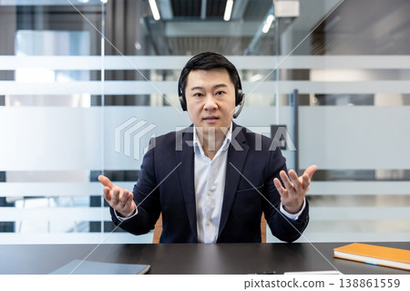 Asian businessman in a suit wearing a headset, engaging in a virtual video conference from an office, discussing business with hand gestures for online communication and remote work Asian businessman in a suit wearing a headset, engaging in a virtual video conference from an office, discussing business with hand gestures for online communication and remote work 138861559