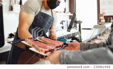 Butcher sells fresh meat to customer in shop with clear hygiene measures 138862448