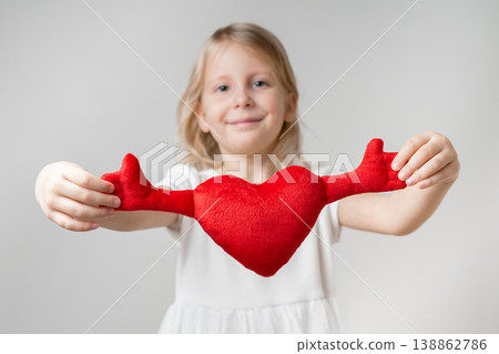 Front view. A cute little girl is holding a soft toy in the shape of a heart Front view. A cute little girl is holding a soft toy in the shape of a heart 138862786