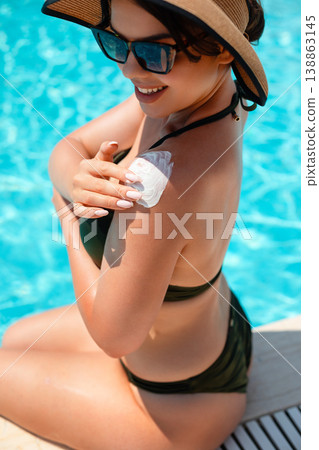 Attractive Woman Applying Sunscreen On Shoulder Sitting By Pool Edge In Summer Sunlight. Skincare And Sun Protection Concept With Healthy Skin Glow Attractive Woman Applying Sunscreen On Shoulder Sitting By Pool Edge In Summer Sunlight. Skincare And Sun Protection Concept With Healthy Skin Glow 138863145