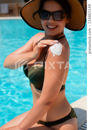 Smiling woman in black bikini and straw hat sitting by pool applying sunscreen on shoulder enjoying bright sunlight and happy summer vacation mood with blue water 138863149