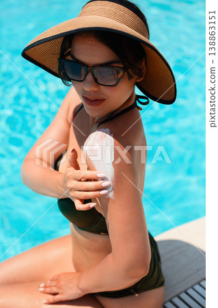 Woman in black bikini and straw hat sitting by pool applying sunscreen on shoulder holding bottle enjoying bright sunlight and relaxed summer vacation atmosphere Woman in black bikini and straw hat sitting by pool applying sunscreen on shoulder holding bottle enjoying bright sunlight and relaxed summer vacation atmosphere 138863151