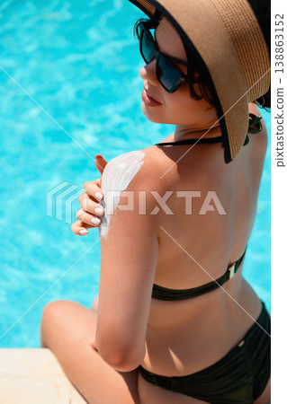 Woman in black bikini and straw hat sitting by pool from back angle applying sunscreen on shoulder enjoying bright sunlight and relaxed summer vacation mood Woman in black bikini and straw hat sitting by pool from back angle applying sunscreen on shoulder enjoying bright sunlight and relaxed summer vacation mood 138863152