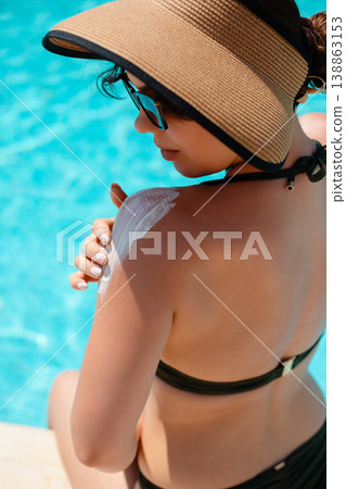 Woman in black bikini and sunglasses sitting by pool from back view applying sunscreen on shoulder enjoying warm sunlight and calm summer atmosphere 138863153