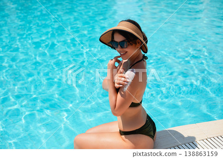 Beautiful Woman Applying Sunscreen Sitting By Poolside In Bright Summer Sunlight With Clear Blue Water. Skincare Routine And Sun Protection Concept Beautiful Woman Applying Sunscreen Sitting By Poolside In Bright Summer Sunlight With Clear Blue Water. Skincare Routine And Sun Protection Concept 138863159