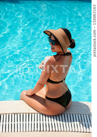Beautiful Woman Sitting By Poolside Relaxing After Applying Sunscreen In Bright Summer Sunlight. Skincare And Healthy Lifestyle Concept Beautiful Woman Sitting By Poolside Relaxing After Applying Sunscreen In Bright Summer Sunlight. Skincare And Healthy Lifestyle Concept 138863163