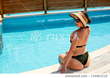 Attractive Woman Sitting by Pool with Sunscreen on Back in Summer Sun. Skincare Routine Concept Attractive Woman Sitting by Pool with Sunscreen on Back in Summer Sun. Skincare Routine Concept 138863228