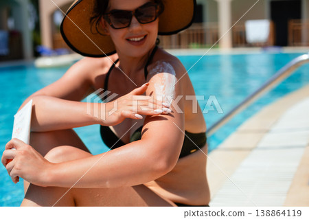Woman Smiling While Applying Sunscreen On Shoulder Sitting By Poolside In Summer Sunlight. Skincare And Sun Protection Concept 138864119