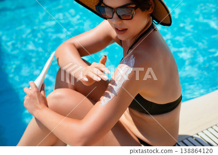 Woman Sitting Poolside Applying Sunscreen On Shoulder With Relaxed Summer Pose And Bright Blue Water Background Woman Sitting Poolside Applying Sunscreen On Shoulder With Relaxed Summer Pose And Bright Blue Water Background 138864120