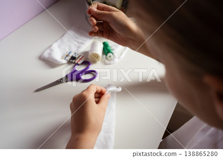 close up view girl sits at a white table sewing white fabric with a needle diy 138865280
