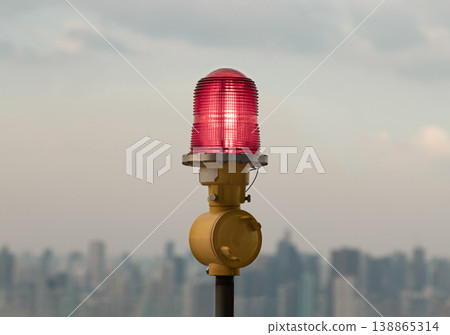 Against a hazy, metropolitan horizon, a singular red obstruction light or aircraft warning light stands as a silent sentinel. The beacon, encased in a crimson, ribbed glass housing, glows with a soft Against a hazy, metropolitan horizon, a singular red obstruction light or aircraft warning light stands as a silent sentinel. The beacon, encased in a crimson, ribbed glass housing, glows with a soft 138865314