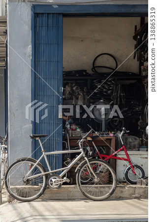 A silver bicycle stands in profile, its metallic frame gleaming against a weathered blue shop door. Beside it, a petite crimson companion offers a vibrant contrast to the workshop industrial shadows. A silver bicycle stands in profile, its metallic frame gleaming against a weathered blue shop door. Beside it, a petite crimson companion offers a vibrant contrast to the workshop industrial shadows. 138865319