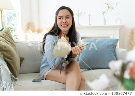 Joyful Asia woman Relaxing on Sofa with book diary. A young woman finds peace and tranquility as she relaxes on a comfortable sofa, immersed in the captivating world of literature. Lifestyle Concept Joyful Asia woman Relaxing on Sofa with book diary. A young woman finds peace and tranquility as she relaxes on a comfortable sofa, immersed in the captivating world of literature. Lifestyle Concept 138867477