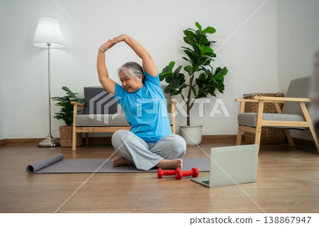 An elderly woman follows an online workout video in her living room, stretching on a yoga mat beside dumbbells. The scene highlights active aging, home fitness, and the use of technology 138867947