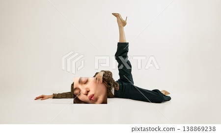 Young woman lying with leg raised holding printed face panel minimal fashion studio portrait. Young woman lying with leg raised holding printed face panel minimal fashion studio portrait. 138869238