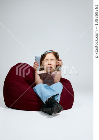 Little girl making funny face while holding laptop with portrait print sitting on bean bag in studio 138869263