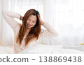 Cheerful Asian woman stretching arms and smiling while waking up on a white bed in a bright bedroom at home. Morning routine, healthy lifestyle, and wellness concept with natural window light. 138869438