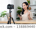 Content Creator in Action: A young content creator, full of energy, broadcasts live from a well-lit studio. With a camera set up and her phone in hand. 138869440