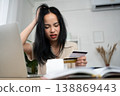 An Asian woman sits indoors holding receipts and a credit card, appearing concerned. The scene reflects financial stress, personal budgeting, and the emotional impact of managing expenses in a home 138869443