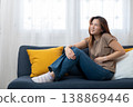 Woman with abdominal pain: A woman is depicted sitting on a sofa, visibly experiencing discomfort and holding her abdomen, suggesting abdominal pain or a stomachache. 138869446