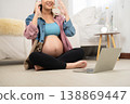 A pregnant woman sits cross-legged on the floor with headphones, engaging with a laptop in a cozy bedroom. The scene highlights modern prenatal lifestyle, digital learning, and maternal wellbeing 138869447