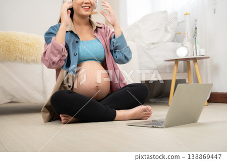 A pregnant woman sits cross-legged on the floor with headphones, engaging with a laptop in a cozy bedroom. The scene highlights modern prenatal lifestyle, digital learning, and maternal wellbeing 138869447