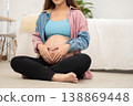 A pregnant woman sits cross-legged in a cozy room, gently forming a heart shape over her belly. The scene captures maternal anticipation, emotional connection, and the warmth of prenatal love 138869448