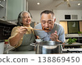 Two senior adults enjoy cooking together in a cozy kitchen. One offers a spoonful of food while the other leans in to smell, capturing warmth, sensory joy, and togetherness in a home lifestyle setting 138869450
