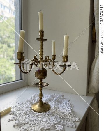 Vintage brass candelabrum with unlit candles on windowsill, white crochet doily background 138870212