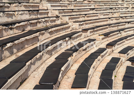 Stone amphitheater seating with rows of steps and black cushions arranged in curved tiers, outdoor venue for audience and events. Amphitheater seating usage for event venue, performance space Stone amphitheater seating with rows of steps and black cushions arranged in curved tiers, outdoor venue for audience and events. Amphitheater seating usage for event venue, performance space 138871627