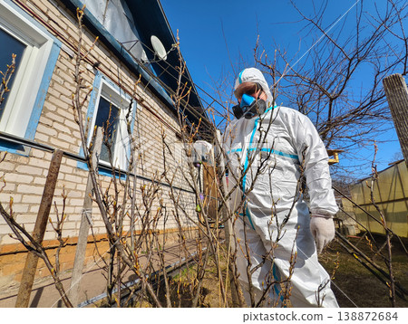 Person wearing protective hazmat suit, respirator mask, and gloves spraying fruit bushes in a spring garden near an old brick house, ensuring chemical safety and plant protection. Person wearing protective hazmat suit, respirator mask, and gloves spraying fruit bushes in a spring garden near an old brick house, ensuring chemical safety and plant protection. 138872684