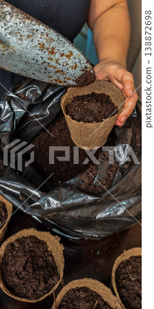 Woman's hands using a gardening trowel to fill an eco-friendly biodegradable pot with dark soil, preparing it for planting vegetable or flower seedlings, promoting sustainable home gardening. Woman's hands using a gardening trowel to fill an eco-friendly biodegradable pot with dark soil, preparing it for planting vegetable or flower seedlings, promoting sustainable home gardening. 138872698