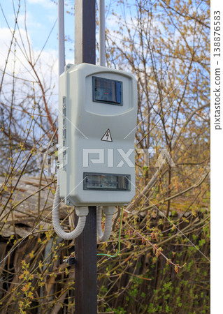 Outdoor electric meter box mounted on a metal pole in a rural area. Plastic electrical cabinet with circuit breakers and high voltage sign. 138876583