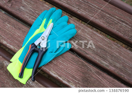 Bright green and blue garden gloves with a professional pruner on a weathered wooden bench. Essential horticulture tools for spring plant maintenance Bright green and blue garden gloves with a professional pruner on a weathered wooden bench. Essential horticulture tools for spring plant maintenance 138876590