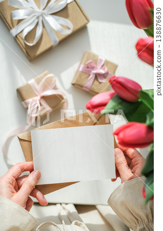 Vertical shot of woman's hands holding a blank white card over table with red tulips and craft gifts. Greeting card mockup Vertical shot of woman's hands holding a blank white card over table with red tulips and craft gifts. Greeting card mockup 138876630