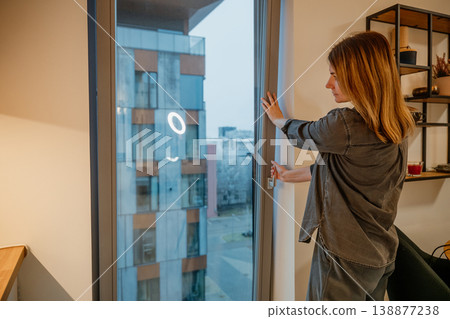 Woman opens window in modern apartment during overcast day in urban setting Woman opens window in modern apartment during overcast day in urban setting 138877238
