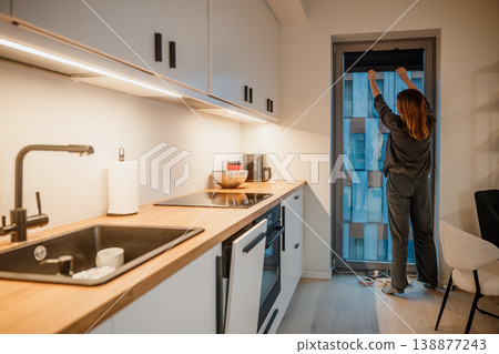 Woman closing blinds in modern kitchen during evening time in an urban apartment Woman closing blinds in modern kitchen during evening time in an urban apartment 138877243