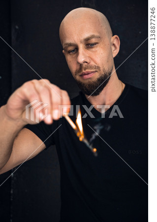 Man holds a match and watches it ignite while standing indoors near a black background 138877246